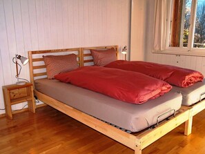 Una cama de madera con dos almohadas y una colcha, una mesita de noche con una lámpara, y una ventana con cortinas.
