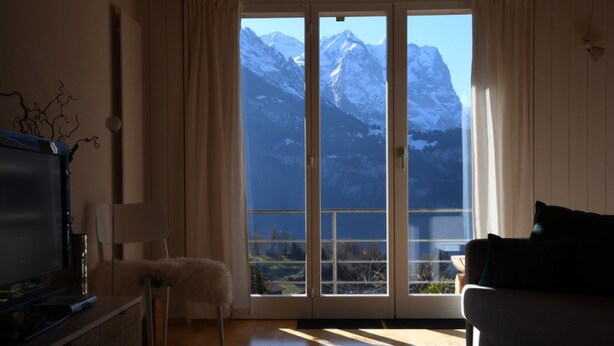 A room with a television, a chair, a sofa, and a view of snow-capped mountains through a sliding door. - HasliRent, Wandelhorn 2, (Hasliberg Reuti), by Interhome (Hasliberg)