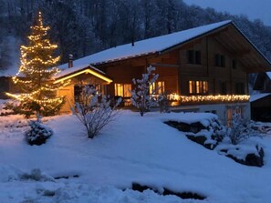 Eine Holzhütte mit schneebedecktem Dach, umgeben von verschneitem Gelände und Bäumen.