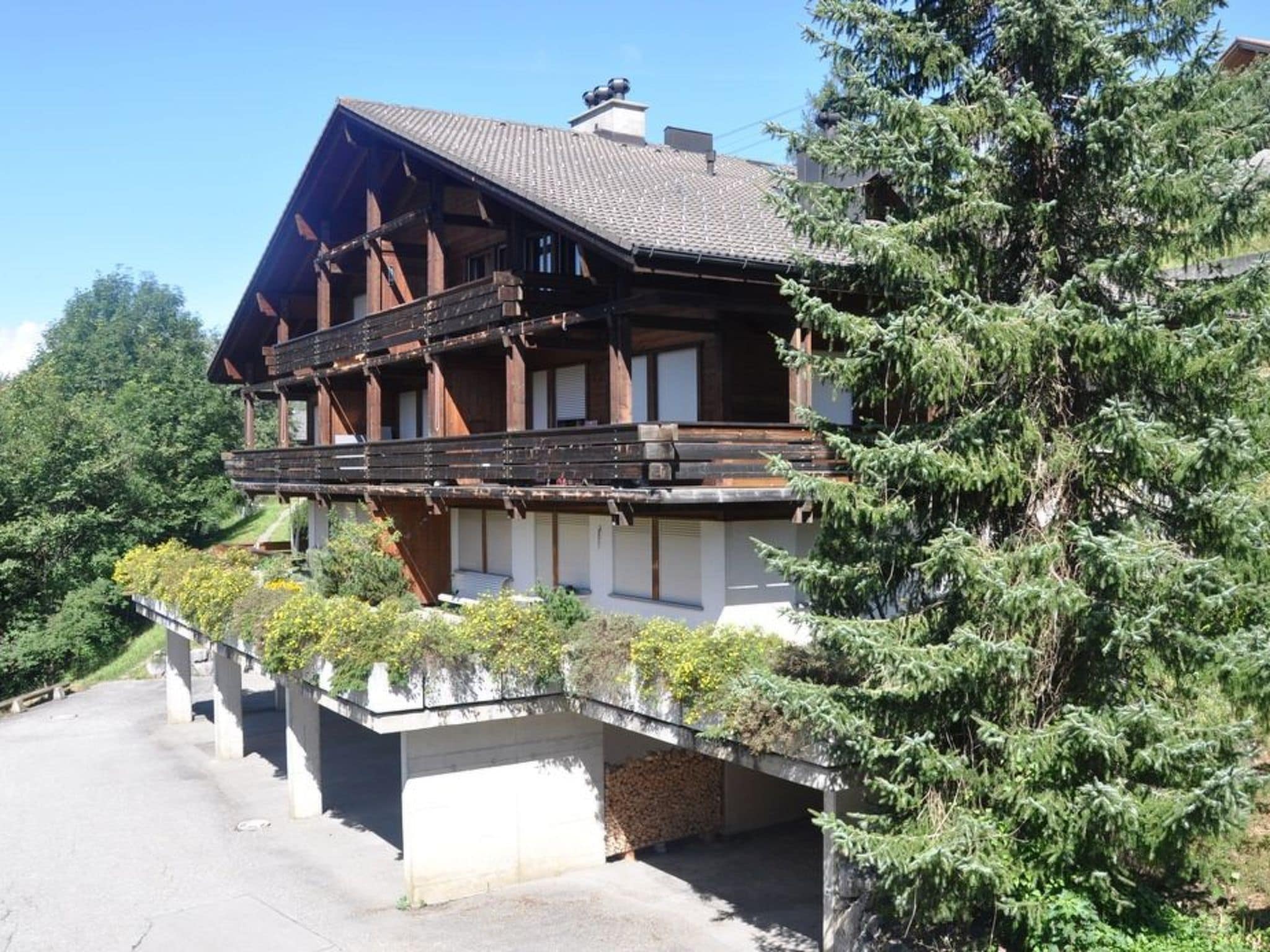 A two-story chalet with a balcony, surrounded by greenery and a clear sky.