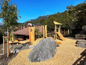 Ein Spielplatz mit Klettergerüsten und einer Rutsche, umgeben von einem Kiesbereich, im Vordergrund ein großer Felsen.