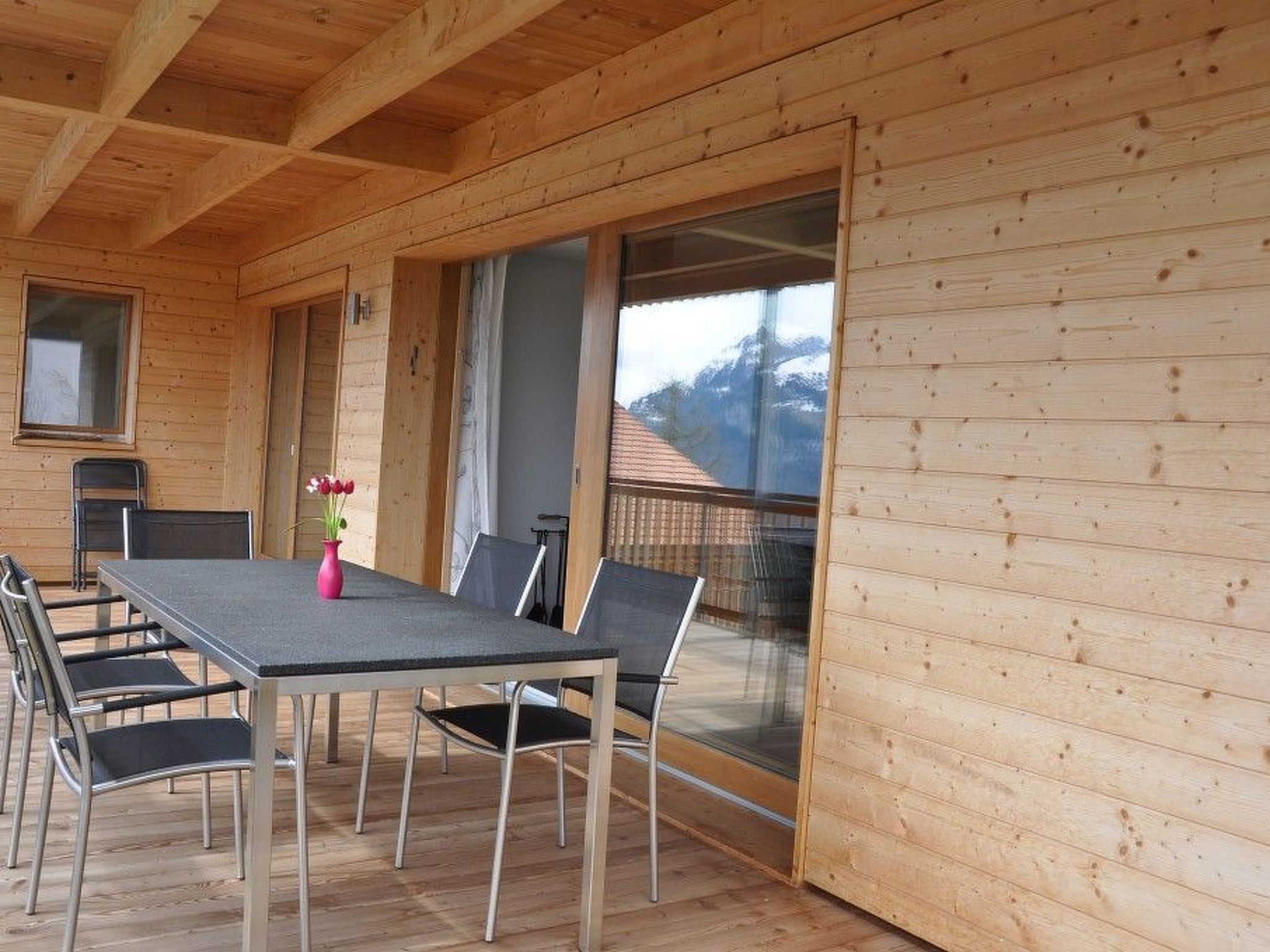 A wooden cabin with a dining area featuring a table and chairs, a window with a view of a mountain, and a balcony with a railing.