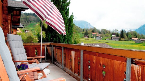 A wooden deck with two lounge chairs and a striped umbrella, overlooking a mountainous landscape. - HasliRent, Diana, (Hasliberg Reuti), by Interhome (Hasliberg)