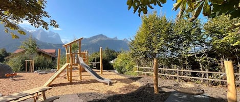 A playground with a slide and climbing structures, surrounded by trees and a mountain backdrop.