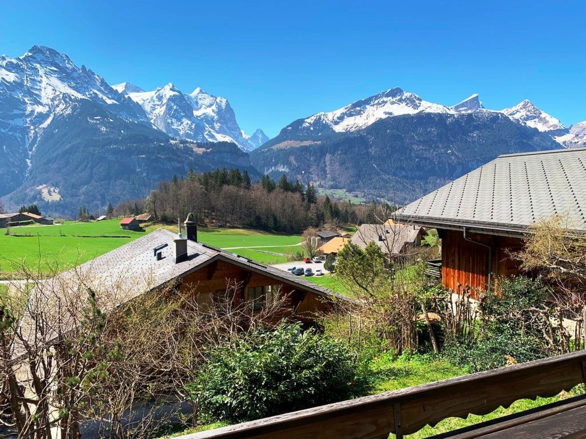 Un paysage de montagne avec des sommets enneigés, une vallée verdoyante et des bâtiments traditionnels en bois.