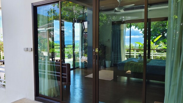 A room with a sliding glass door leading to a balcony, a bed, and a view of a lush landscape. - Deluxe Room No. 2 with Ocean View (Sakhu)