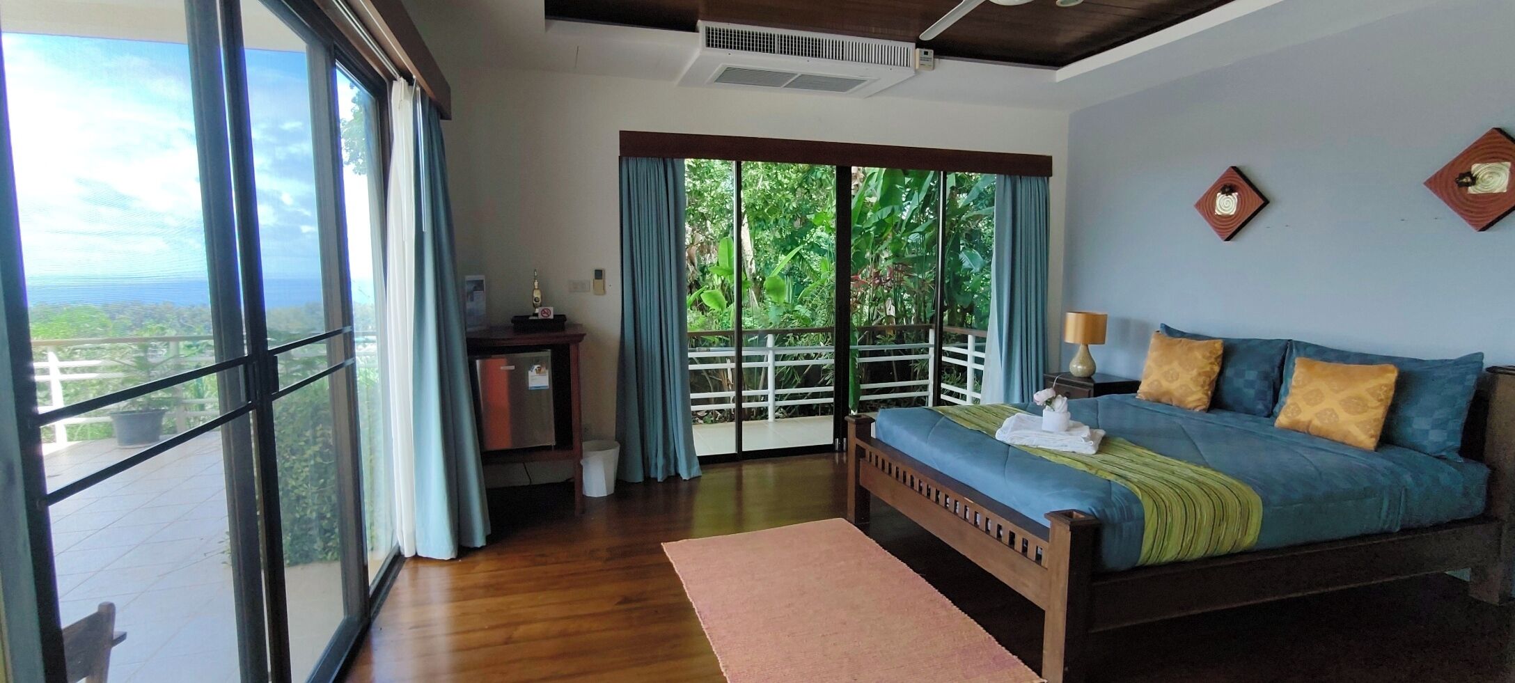 A bedroom with a large sliding glass door leading to a balcony, a bed with blue bedding, a nightstand, and a wall-mounted air conditioning unit.