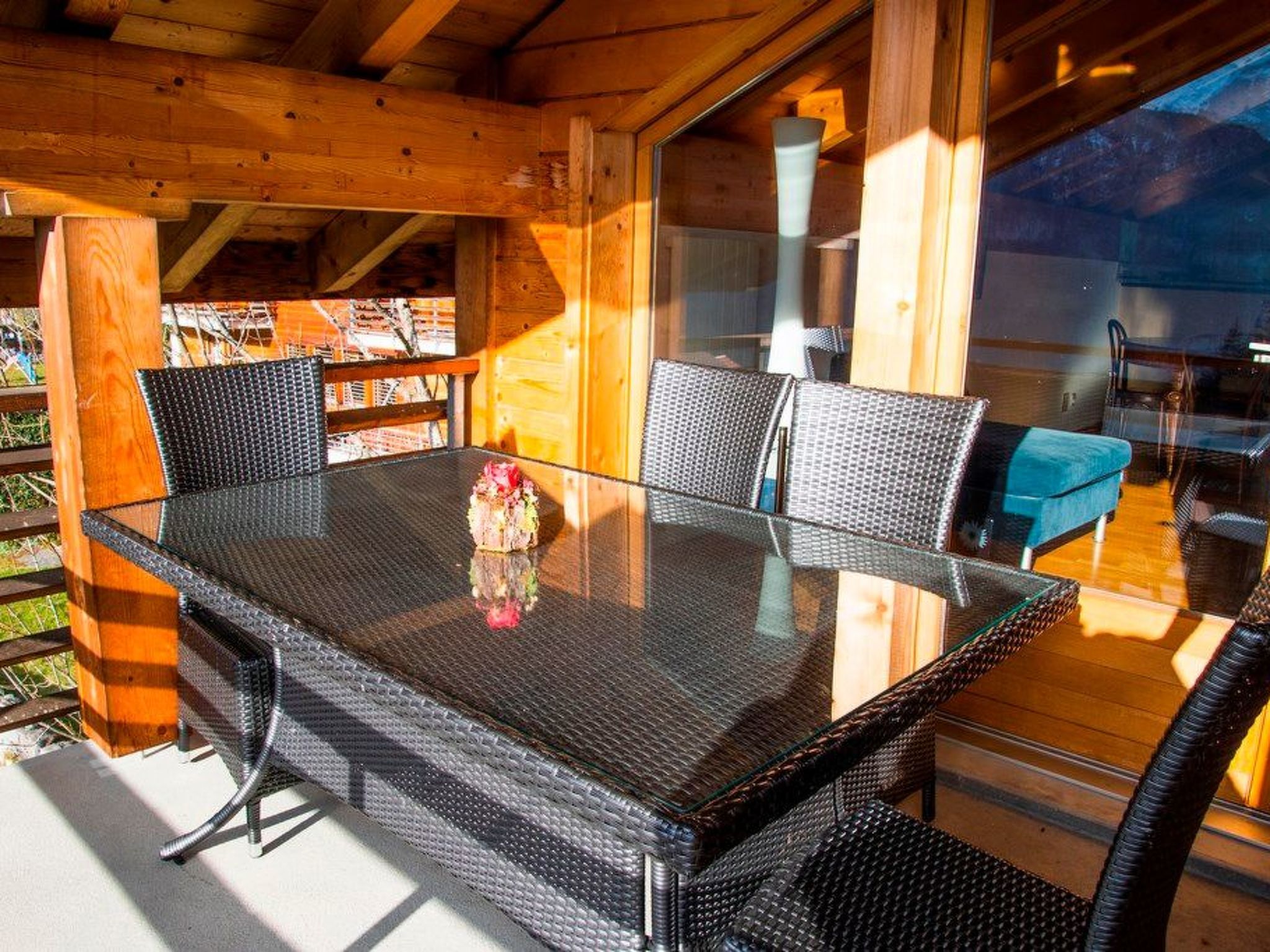 An outdoor dining area with a glass table surrounded by wicker chairs, set against a wooden deck with a view of the surrounding landscape.