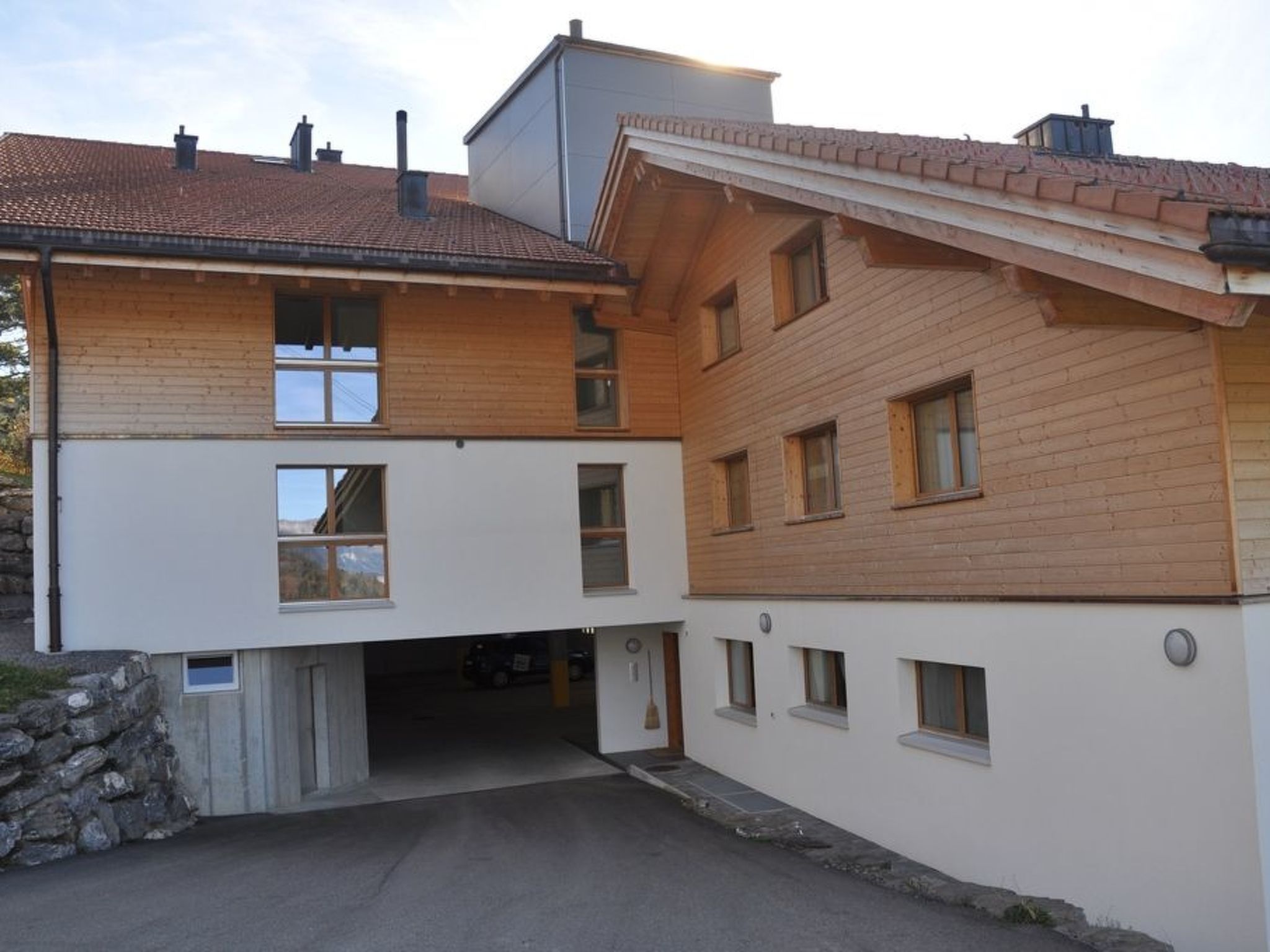 A two-story building with a sloped roof, a garage entrance, and a stone wall on the left.