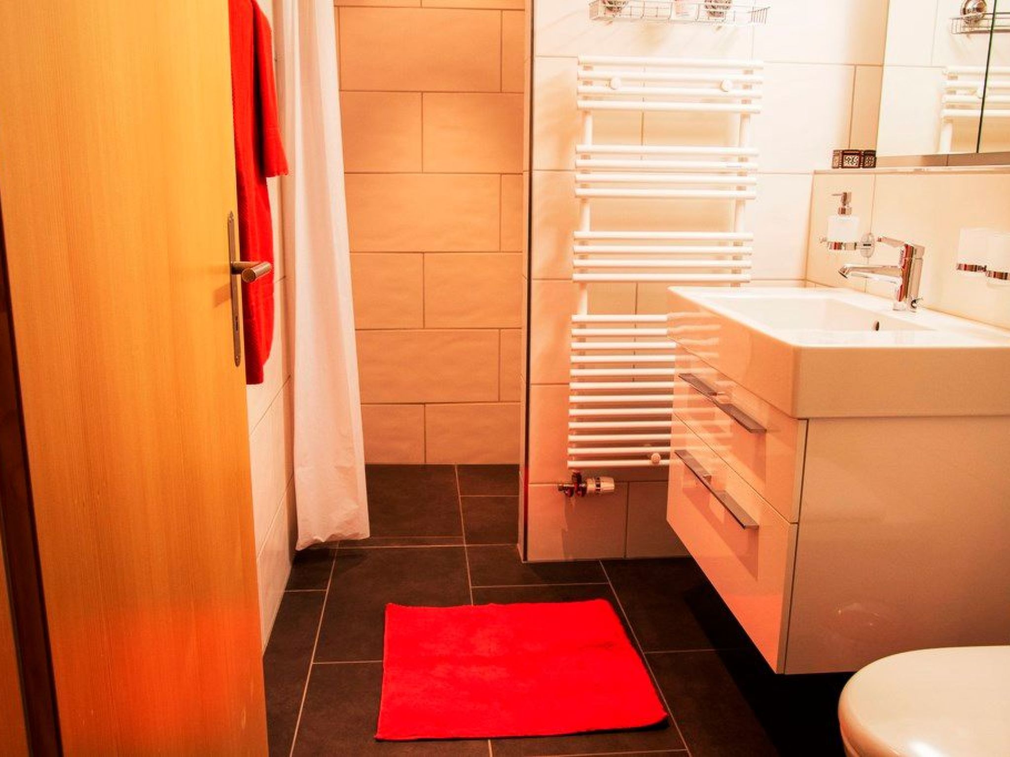 Un baño con lavamanos blanco, un espejo, toallero y felpudo rojo.
