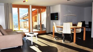 Un salon moderne comprenant un canapé, une table à manger avec des chaises, un téléviseur fixé au mur et de grandes portes-fenêtres coulissantes donnant sur un balcon.