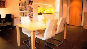 Un espace repas moderne avec une table en bois et des chaises blanches, une bibliothèque, un coin cuisine et une porte fermée.