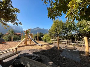 A playground with a slide and climbing structures, surrounded by trees and a mountain backdrop. - HasliRent, Engi West 2, (Hasliberg Reuti), by Interhome (Hasliberg)