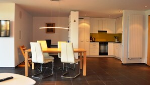 Une cuisine moderne avec des meubles blancs, un coin repas avec une table en bois et des chaises blanches, ainsi qu’une petite table blanche avec une télécommande.