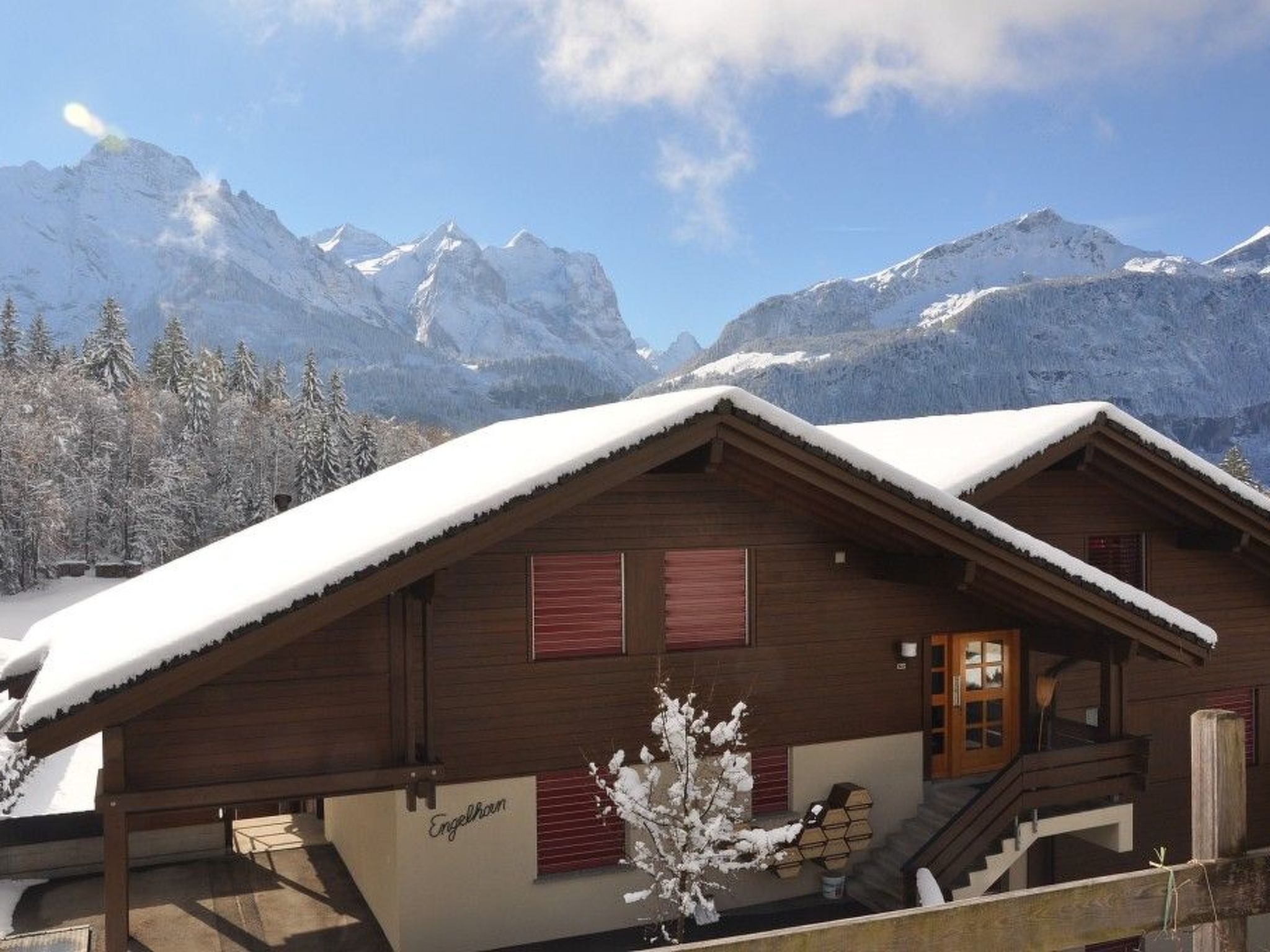A wooden chalet with a snow-covered roof, a wooden staircase, and a view of snow-capped mountains in the background.