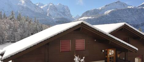 A wooden chalet with a snow-covered roof, a wooden staircase, and a view of snow-capped mountains in the background.