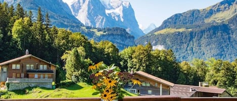 Una mesa de madera con un jarrón blanco lleno de flores, con vistas a un paisaje montañoso con casas y árboles.