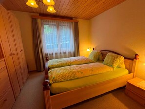 A double bed with patterned bedding, a wooden wardrobe, a window with curtains, and a bedside lamp. - HasliRent, Engelhorn, (Hasliberg Reuti), by Interhome (Hasliberg)