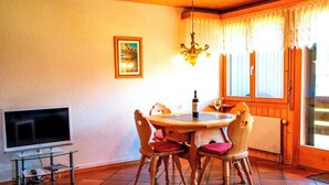 Een eethoek met een ronde tafel, stoelen, een kroonluchter, een televisie op een standaard en een raam met gordijnen.
