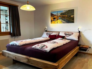 A bedroom with a wooden bed frame, a large window with curtains, a painting on the wall, and a bedside table with a lamp. - HasliRent, Holiboimi, (Hasliberg Wasserwendi), by Interhome (Hasliberg)