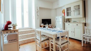 Une cuisine lumineuse avec des meubles blancs, une table à manger avec des chaises, un téléviseur et un réfrigérateur.