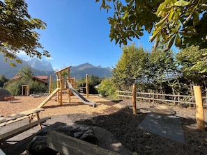 A playground with a slide and climbing structures, surrounded by trees and a mountain backdrop.