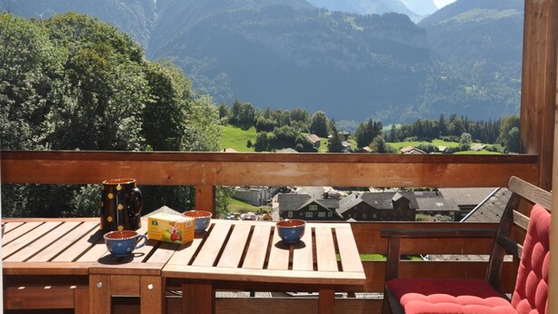 Eine Holzterrasse mit Tisch und Stühlen, mit Blick auf eine gebirgige Landschaft mit Häusern und Grünflächen.