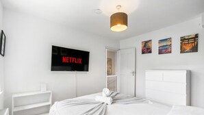 A hotel room with a bed, a TV mounted on the wall, a white headboard, and a shelf with a white object. - Simply Hidden Garden Parking By My Getaways (Horsham)