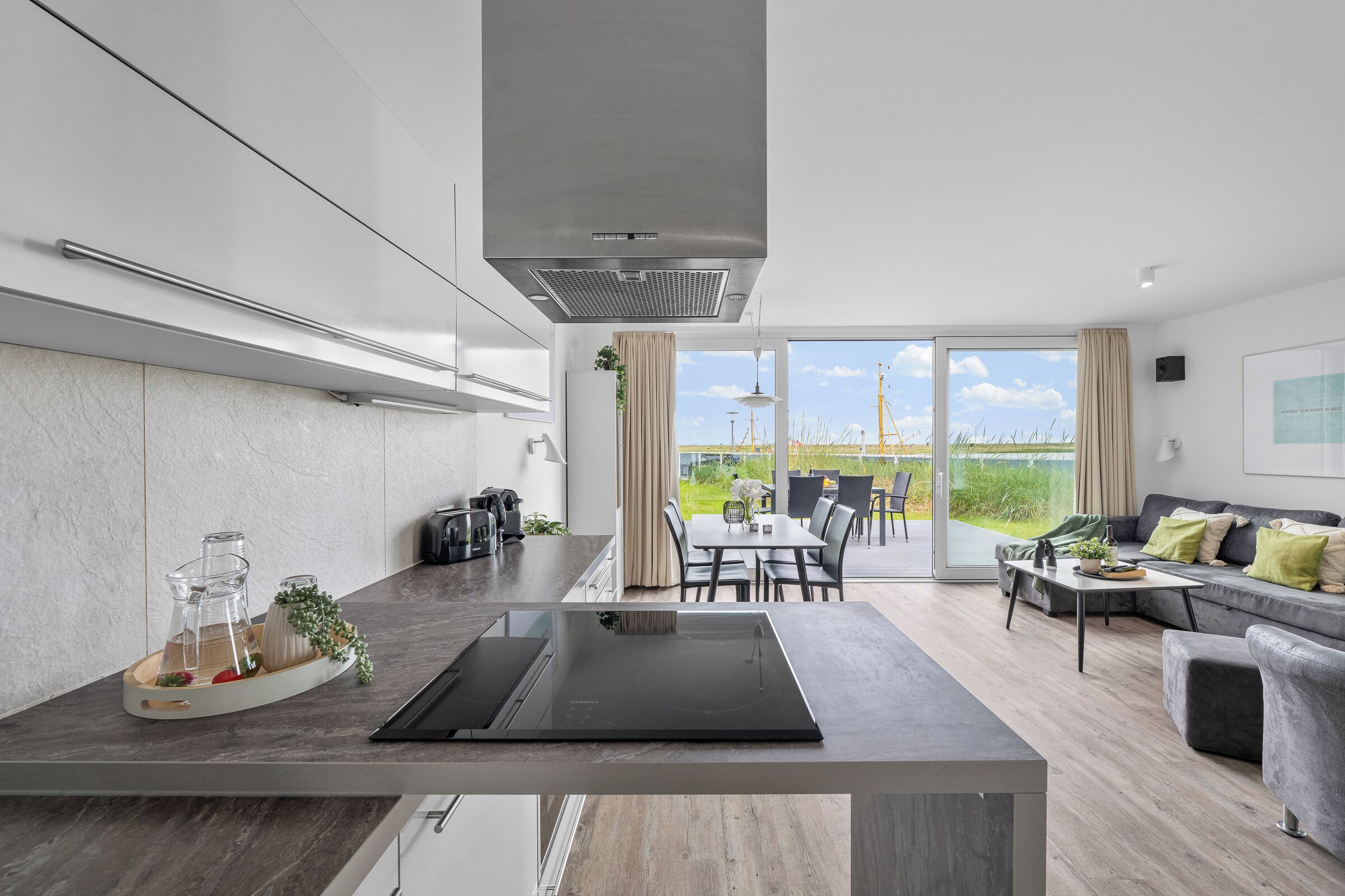 Une cuisine moderne dotée d’un grand îlot central, un coin repas avec une table et des chaises, et un espace de vie avec un canapé et une vue sur l’extérieur.