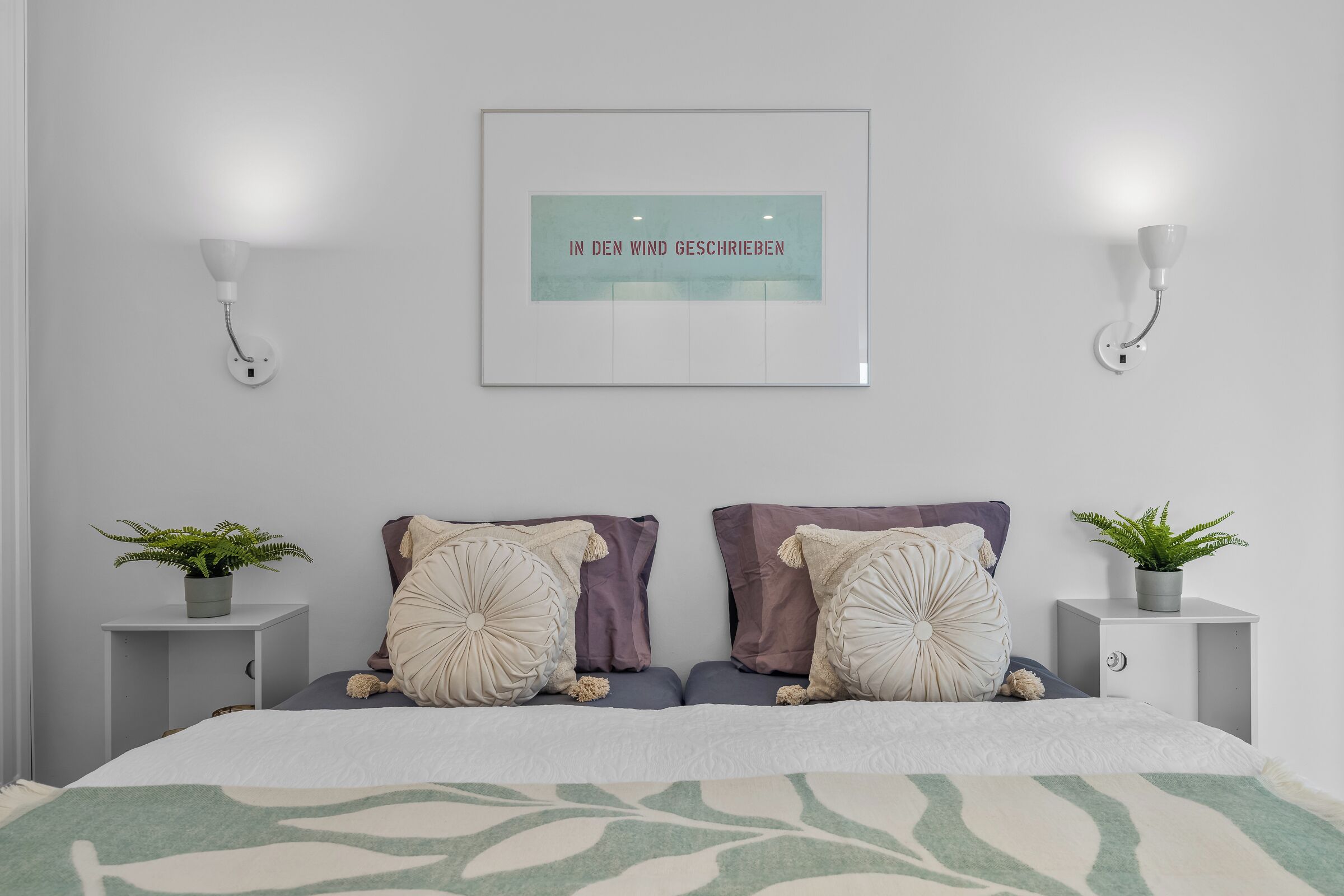 A bedroom with a bed, two bedside tables with plants, a wall-mounted light, and a framed quote.