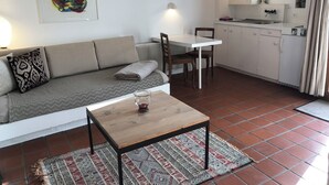 Un salon comprenant un canapé, une table, un tapis, ainsi qu’un coin cuisine avec des placards et une fenêtre.