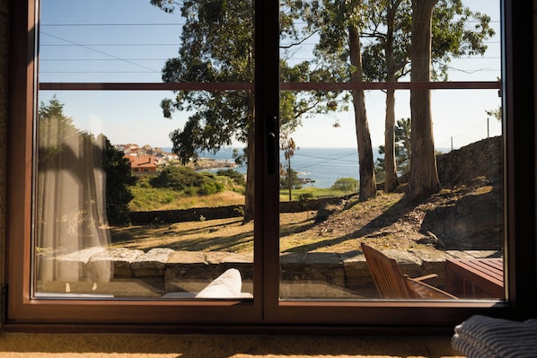 A room with a large window overlooking a coastal landscape, including trees and a distant view of the sea.