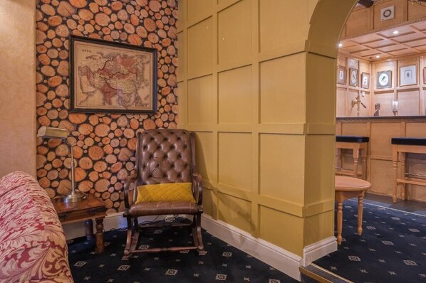 A room with a large map on the wall, a tufted leather chair, a small table with a lamp, and a bar area with wooden paneling and framed pictures.