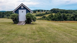 A small blue building with a white door, surrounded by grass and trees, with a hill in the background.