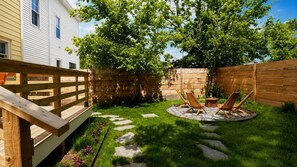 Ein Garten mit einer Holzterrasse, einer runden Feuerstelle aus Stein und zwei Holzstühlen.