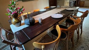 Et spiseområde med et langt bord, stole, hængende lamper, en potteplante og et køkken i baggrunden.