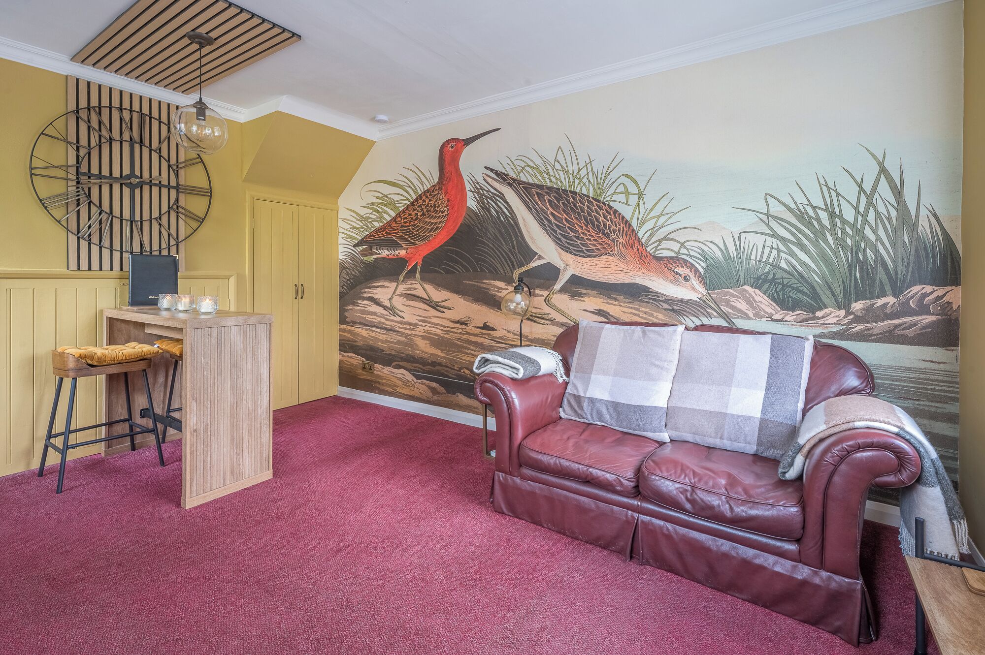 Una habitación con un mural de pájaros, un sofá de cuero rojo, una zona de bar de madera y una alfombra roja.