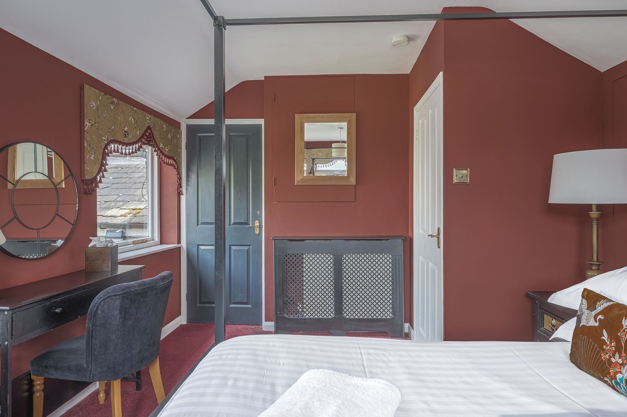 A bedroom with a bed, a desk, a chair, a mirror, a fireplace, and a door.
