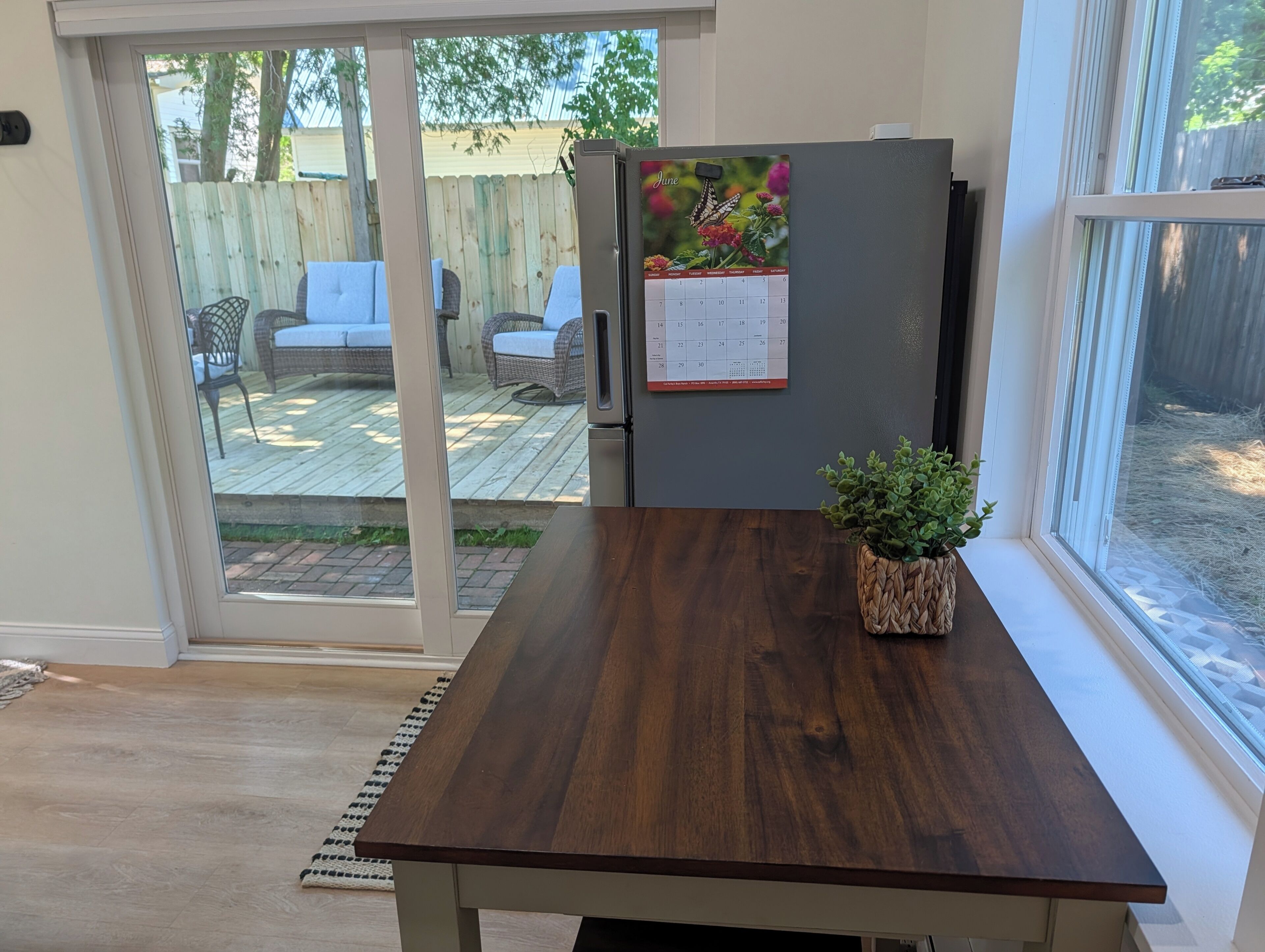Une cuisine avec une table en bois, un réfrigérateur orné d’un calendrier et une porte-fenêtre coulissante donnant sur une terrasse extérieure aménagée avec du mobilier.