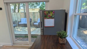 Une cuisine avec une table en bois, un réfrigérateur orné d’un calendrier et une porte-fenêtre coulissante donnant sur une terrasse extérieure aménagée avec du mobilier.