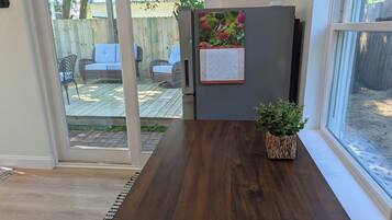 Une cuisine avec une table en bois, un réfrigérateur orné d’un calendrier et une porte-fenêtre coulissante donnant sur une terrasse extérieure aménagée avec du mobilier.