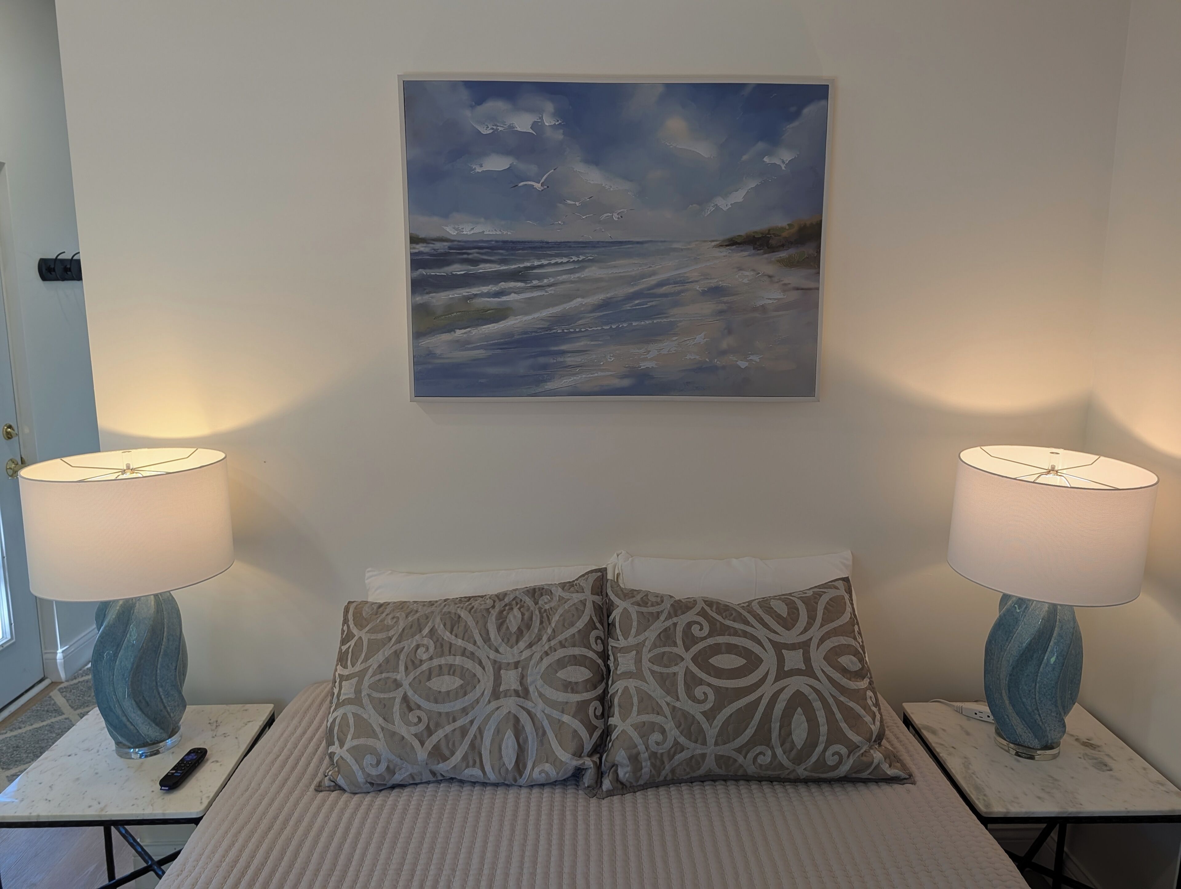 Une chambre à coucher avec un lit, deux lampes de chevet, une télécommande, un coussin à motifs et un tableau représentant une scène de plage.
