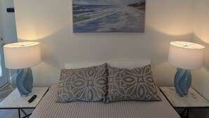 Une chambre à coucher avec un lit, deux lampes de chevet, une télécommande, un coussin à motifs et un tableau représentant une scène de plage.