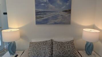 Une chambre à coucher avec un lit, deux lampes de chevet, une télécommande, un coussin à motifs et un tableau représentant une scène de plage.