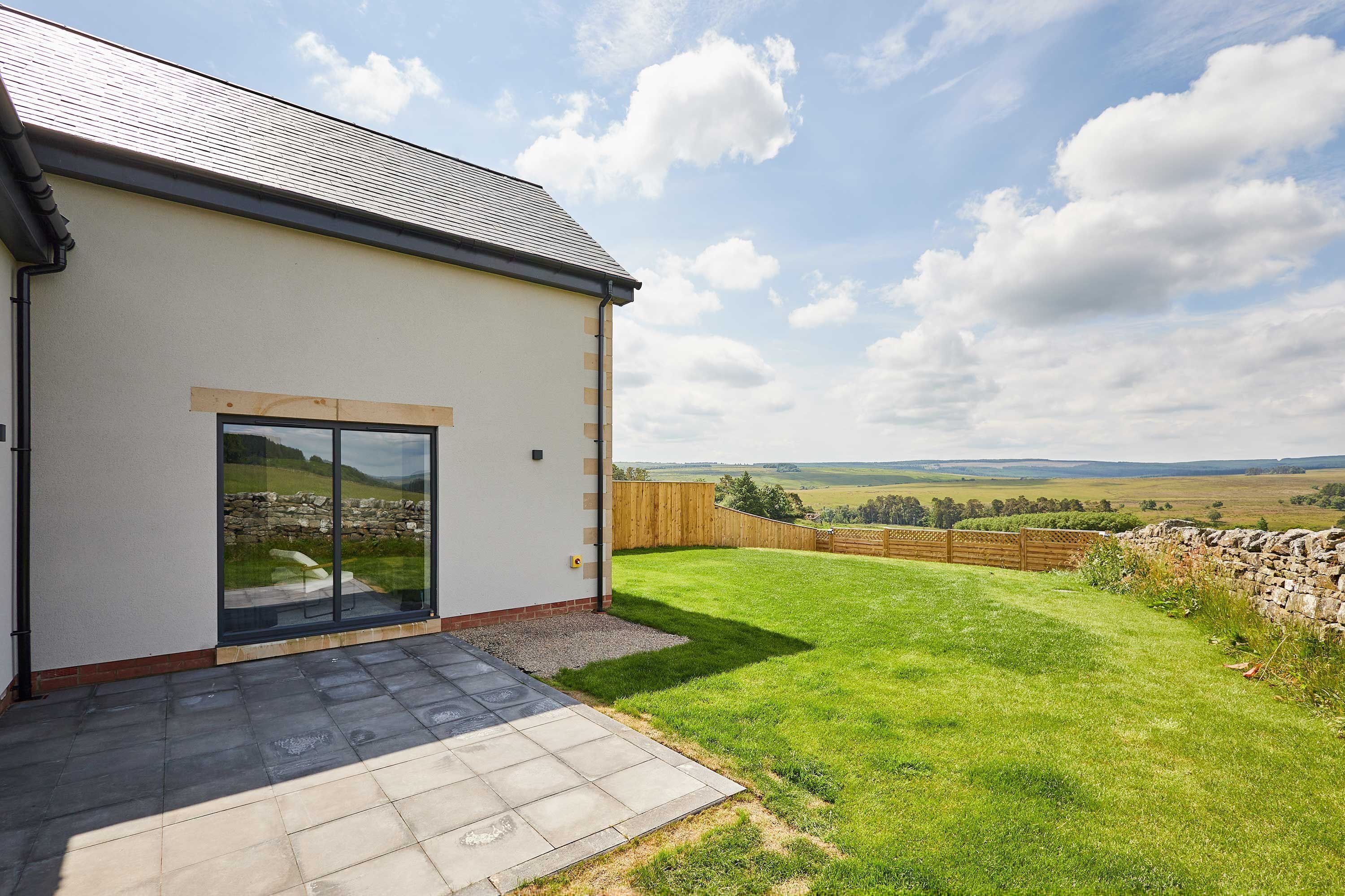 A modern house with a large glass door, a paved patio, and a view of a grassy field.