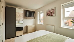 Un dormitorio con cama, cómoda, televisor montado sobre la cómoda, ventana con vistas y puerta que da a otra habitación.