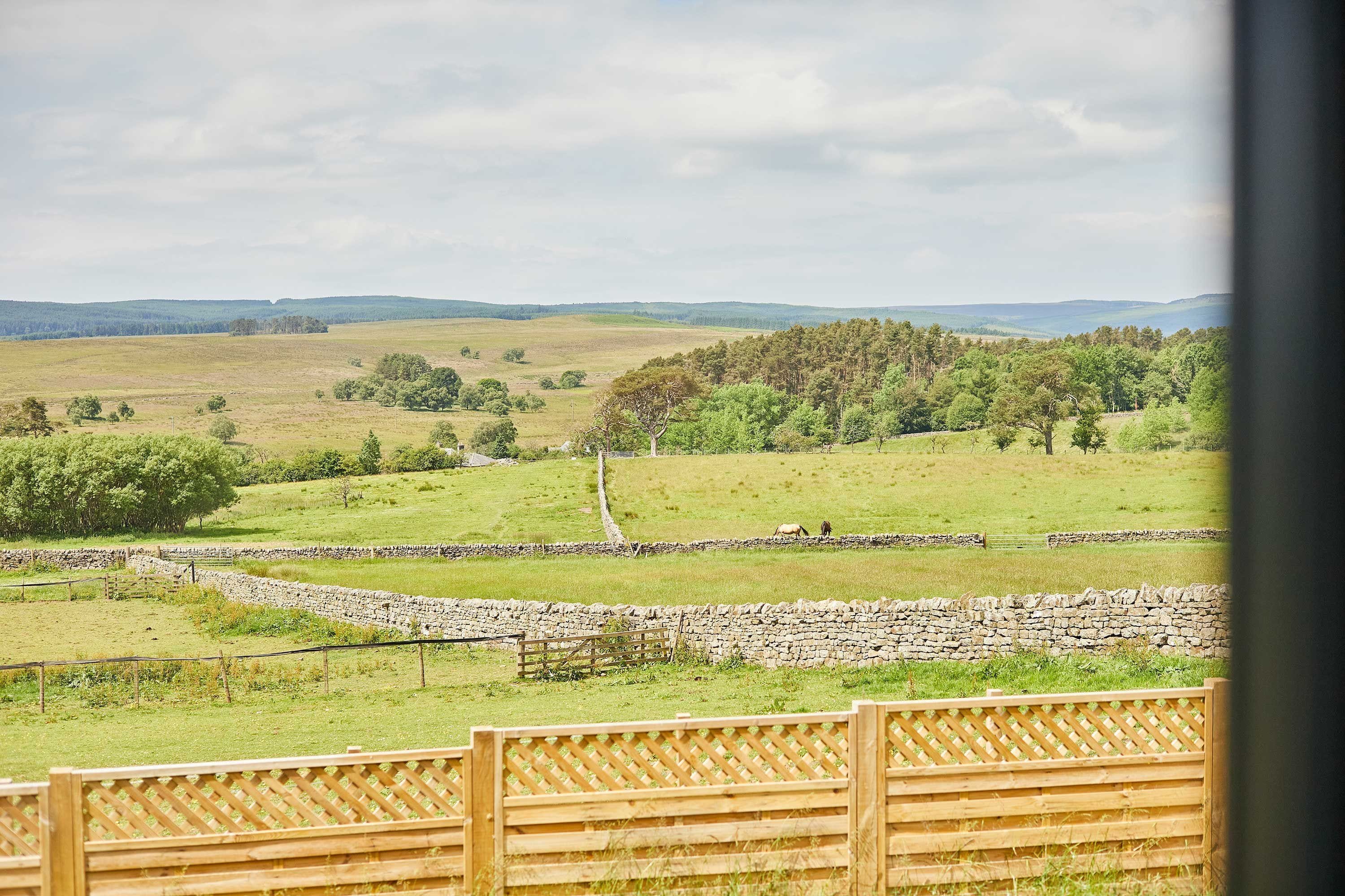 A rural landscape with stone walls, a wooden fence, and a distant view of hills.