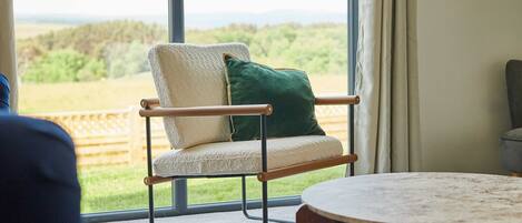 A room with a large window, a chair with a cushion, and a round table.