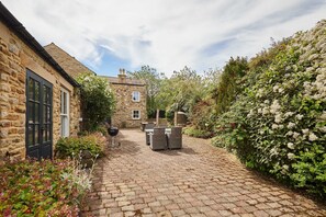 A stone building with a paved courtyard, outdoor furniture, and a garden with flowering plants.