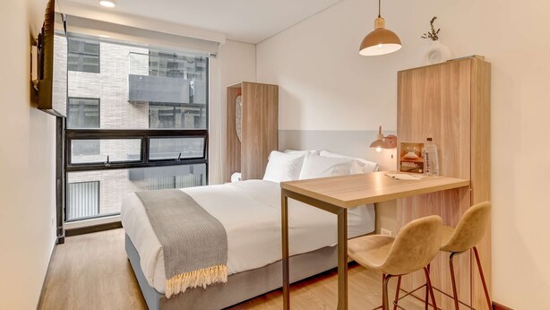 Un dormitorio moderno con cama, mesita de noche y ventana con vista a un edificio.